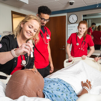 Nursing students work in simulated hospital room; one of them checks the manikin patient’s pupil’s reaction to light.