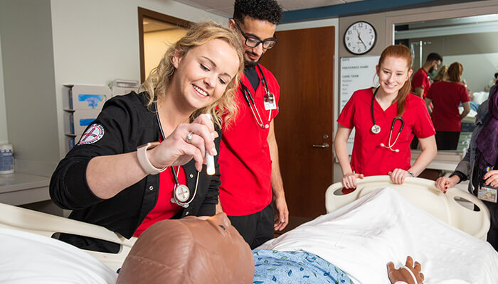 Nursing students work in simulated hospital room; one of them checks the manikin patient’s pupil’s reaction to light.